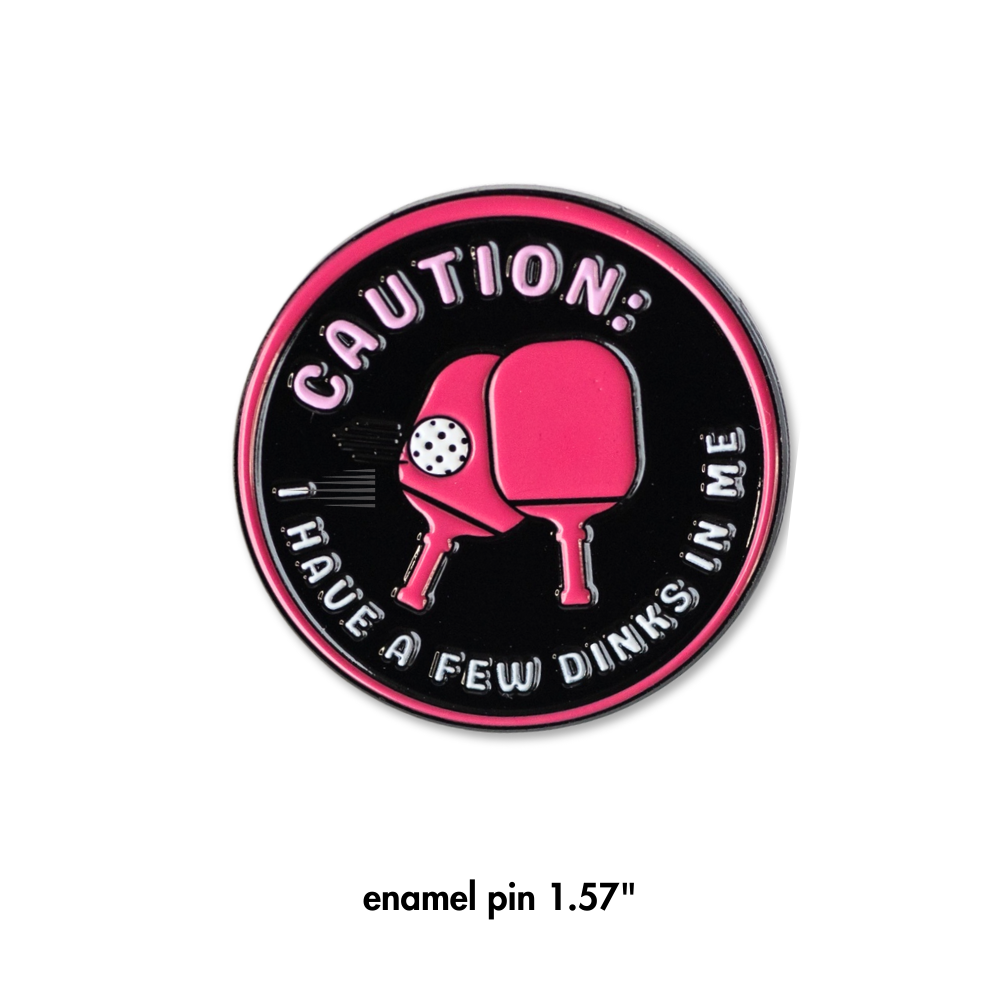 Pink and black enamel pin. Pickleball. Two pink paddles in center. Caution, I have a few dinks in me.