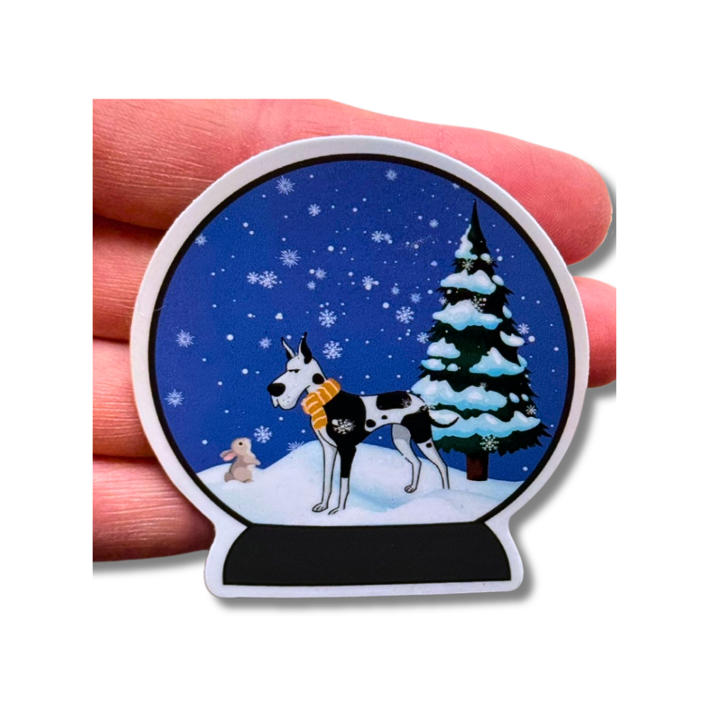 Winter snow globe waterproof sticker - black and white Great Dane dog looking at tiny bunny in the snow. Christmas tree behind them. Shown on top of a woman's hand for size comparison