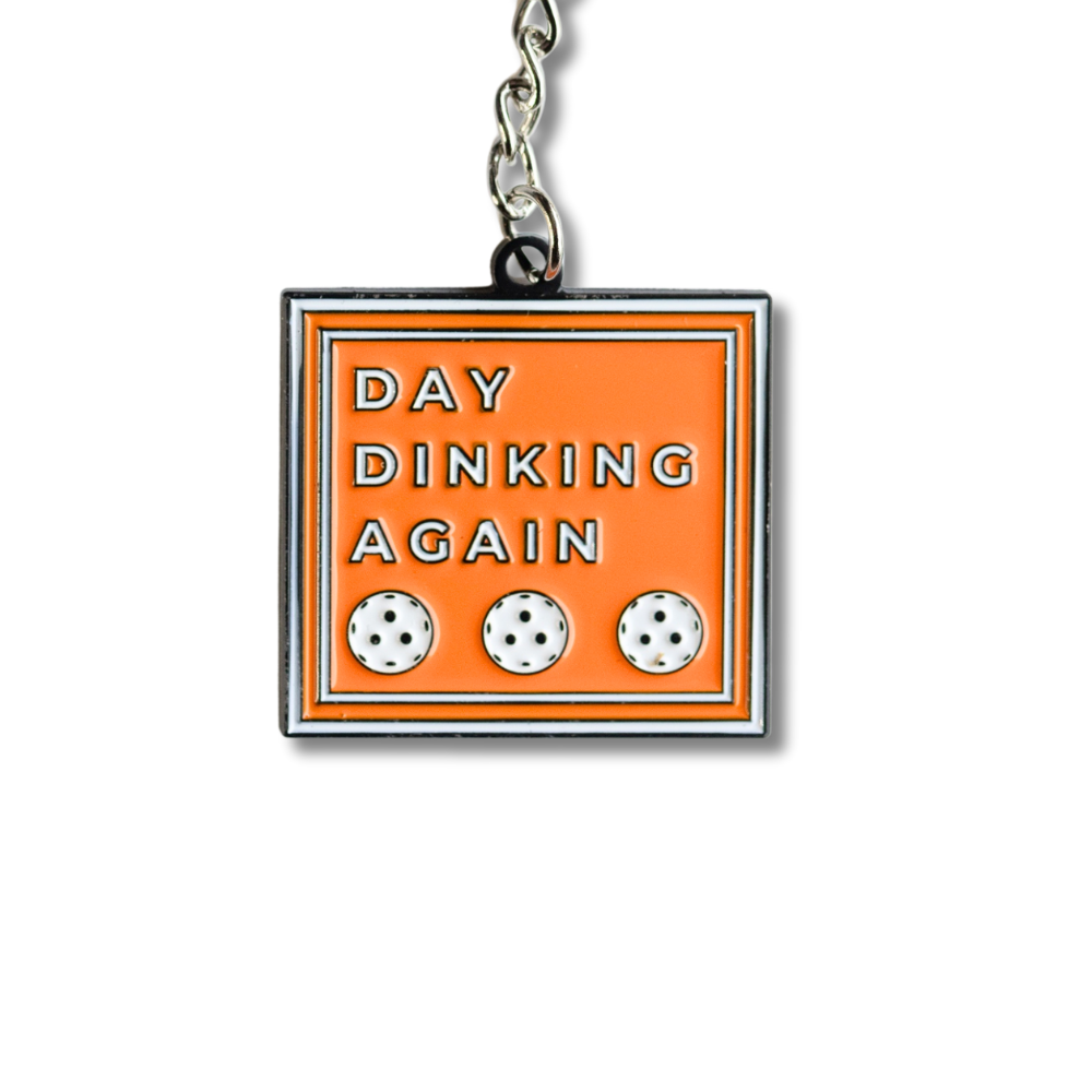 Close up view: Orange square enamel Pickleball keychain. White text with 3 white Pickleballs: Day Dinking Again