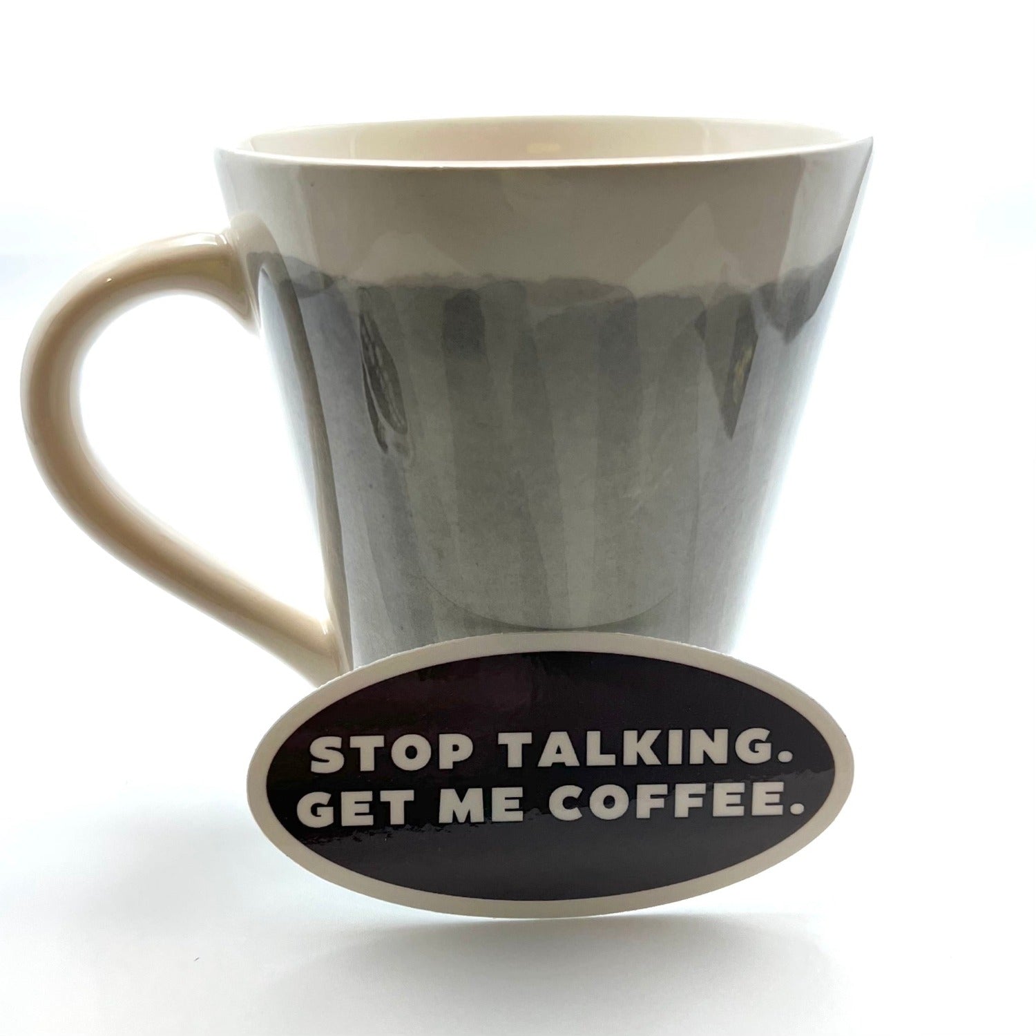 Gray and off-white coffee mug with oval chocolate brown sticker with white font leaning against it, "STOP TALKING. GET ME COFFEE."