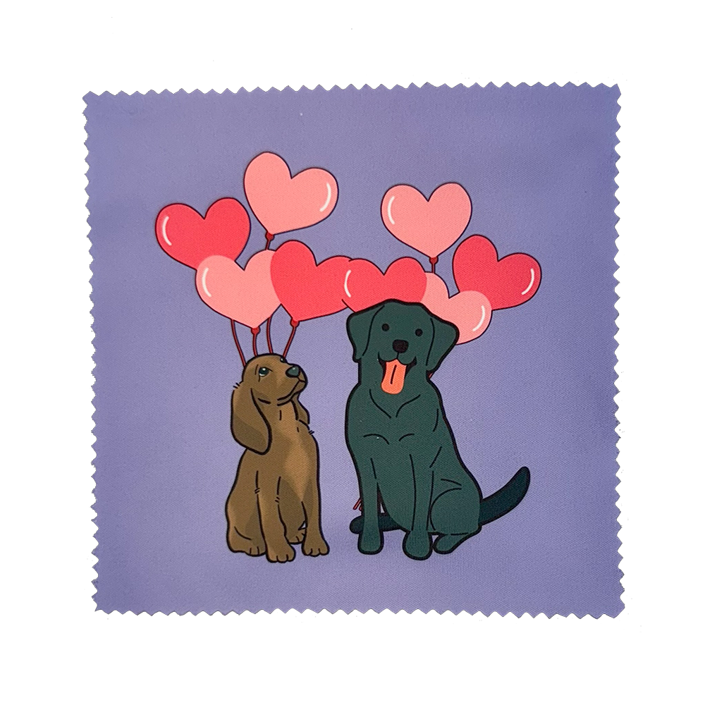 A brown spaniel dog and black lab dog sitting in front of pink heart shaped balloons.  Lavender background.  Lens cloth.