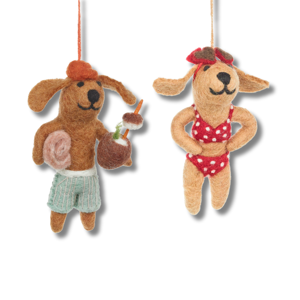 Fair trade felt dog ornaments. The dog on left is wearing swim trunks, holding a towel and holding a coconut cocktail. The pup on the right is wearing a red and white bikini and has red hear glasses on her head.