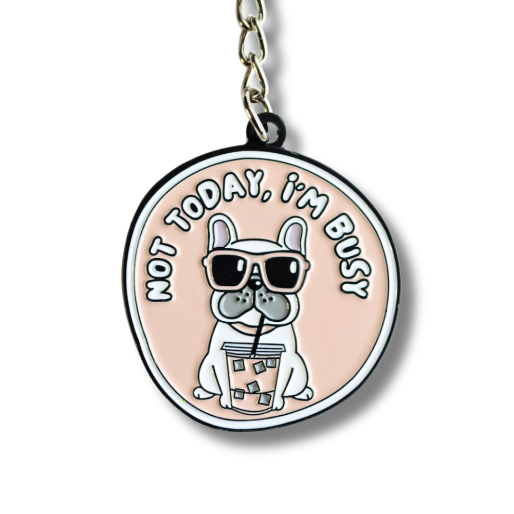 close up of enamel French Bulldog keychain: . Pale pink with white font. White Frenchie drinking bubble tea and looking sassy: not today, I'm busy