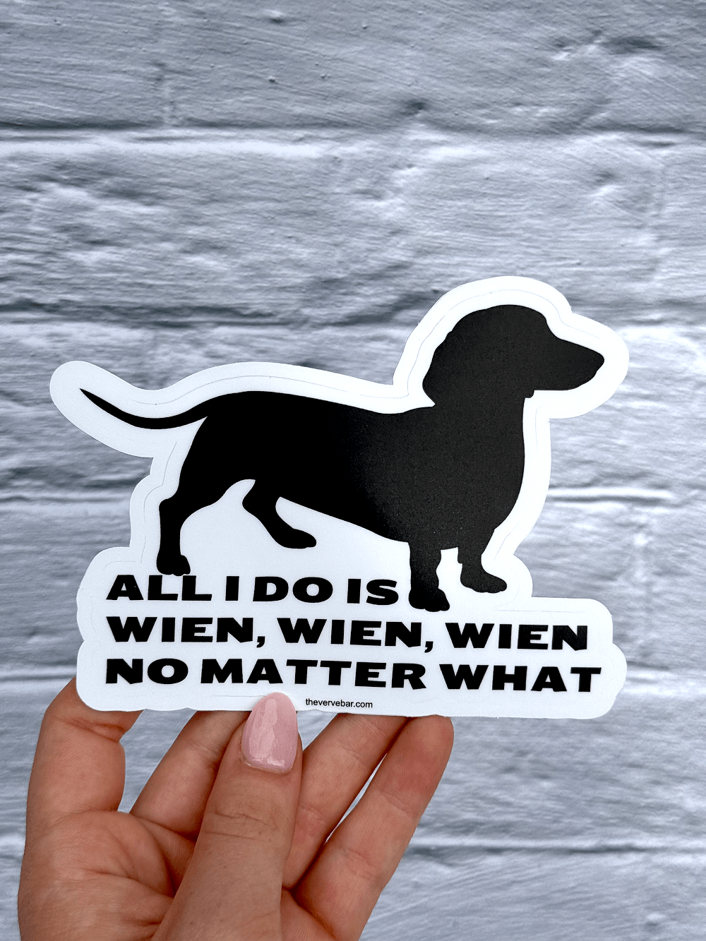 Hand holding large dachshund sticker; dachshund silhouette and text held by a hand against a gray brick background.