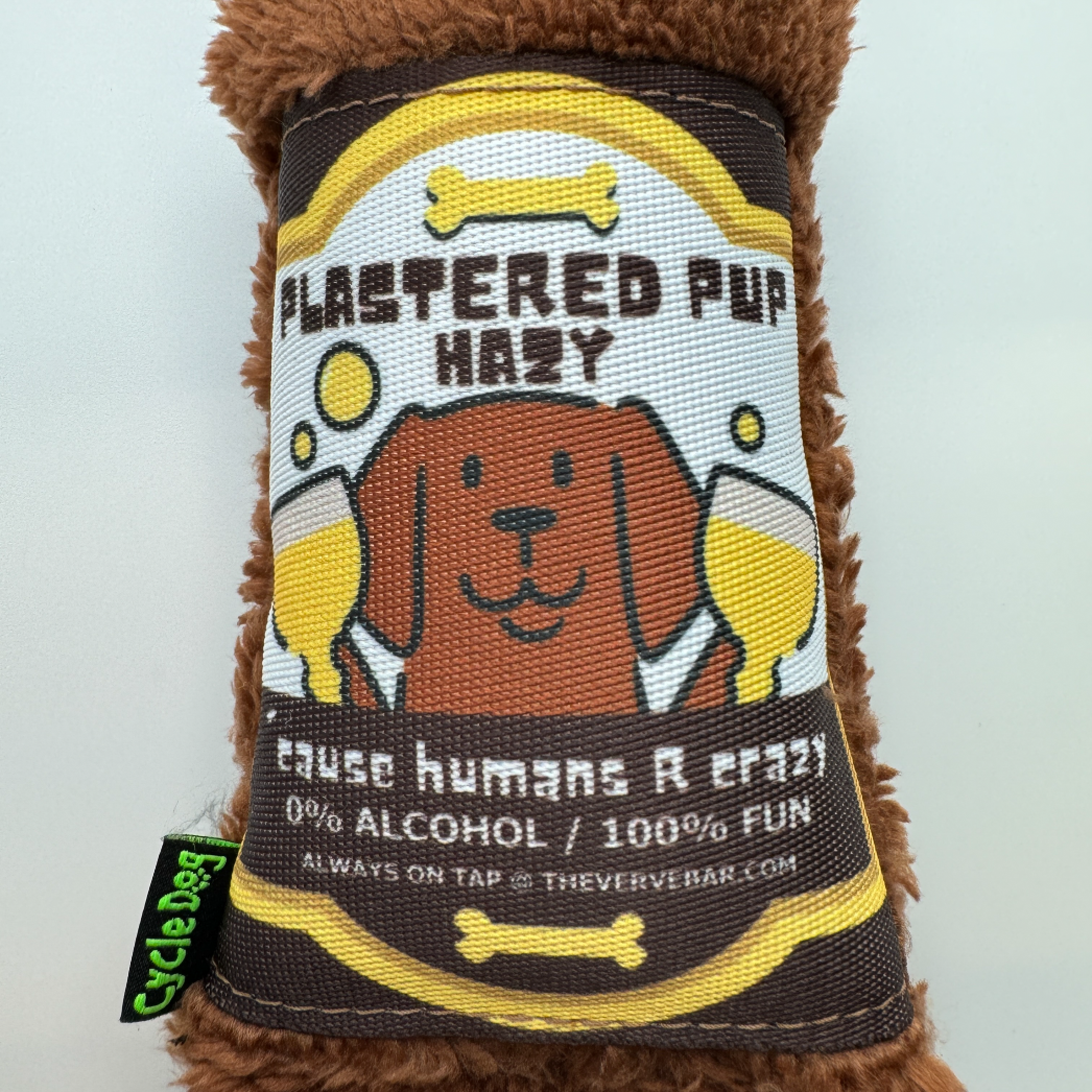 Close of our exclusive funny dog toy beer bottle. The label has a cartoon dog holding two schooners of beer "plastered pup hazy, 'cause humans are crazy."