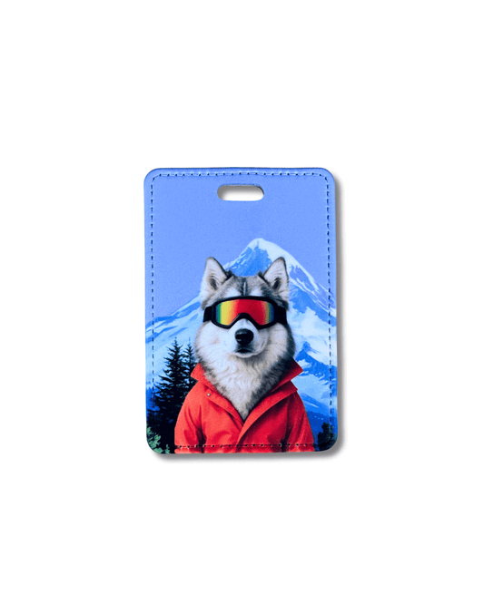 husky ski luggage tag; husky wearing ski goggles and orange jacket with snowy mountain background on blue travel tag