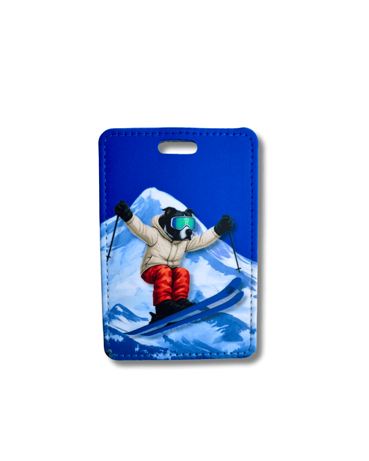 black dog ski luggage tag; wearing ski clothes and skis jumping in air. Mountain in background.