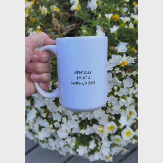 Funny white mug held in front of flowers. Mug has black text: mentally I'm at a swim-up bar