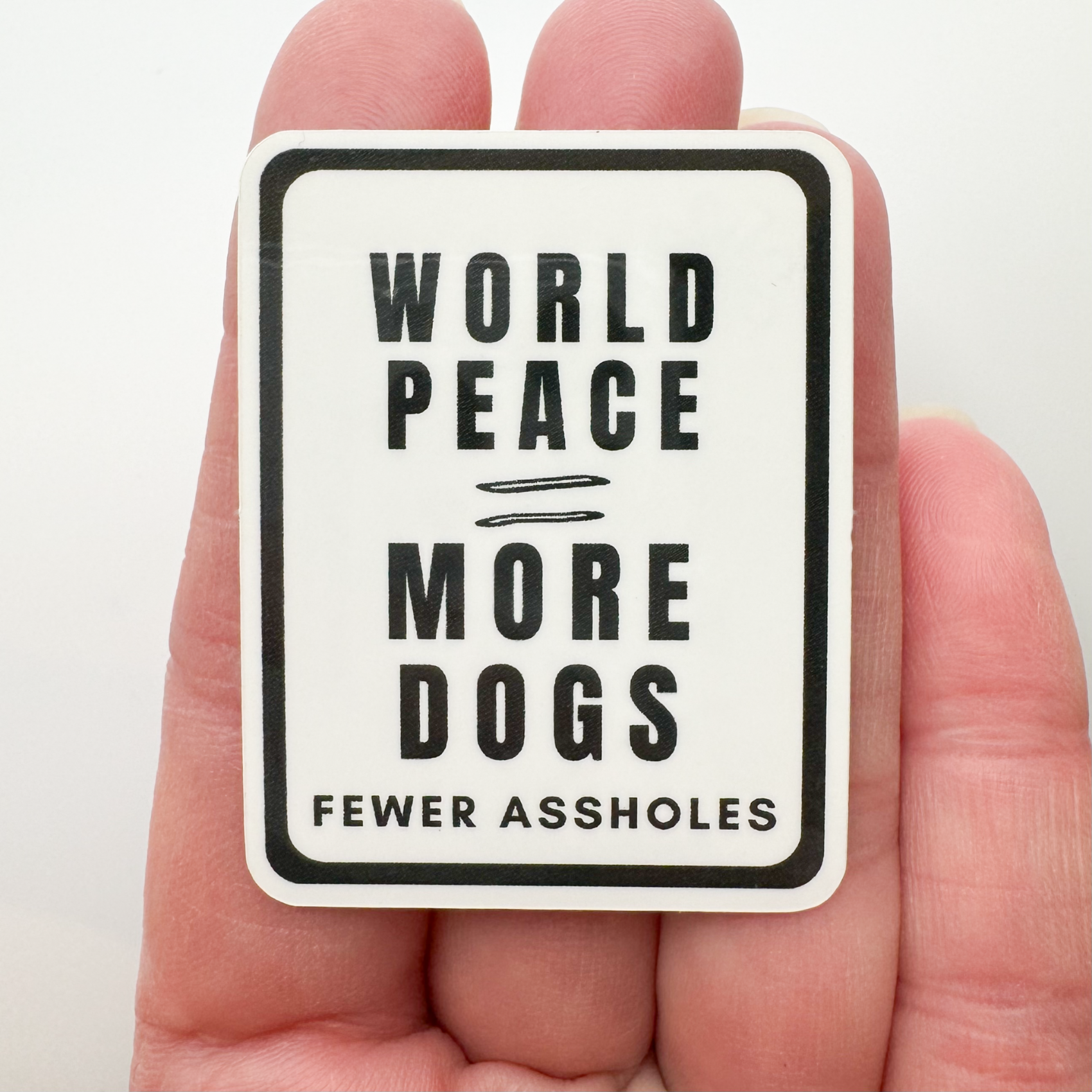 a hand holding the "world peace = more dogs & fewer a-holes"  sticker so you can see the size