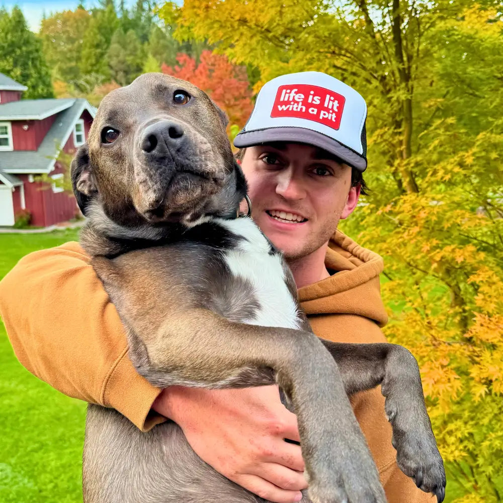 Man wearing black pitbull lover hat and holding black pit bull dog in fall setting. Black trucker hat with red “Life is Lit with a Pit” 